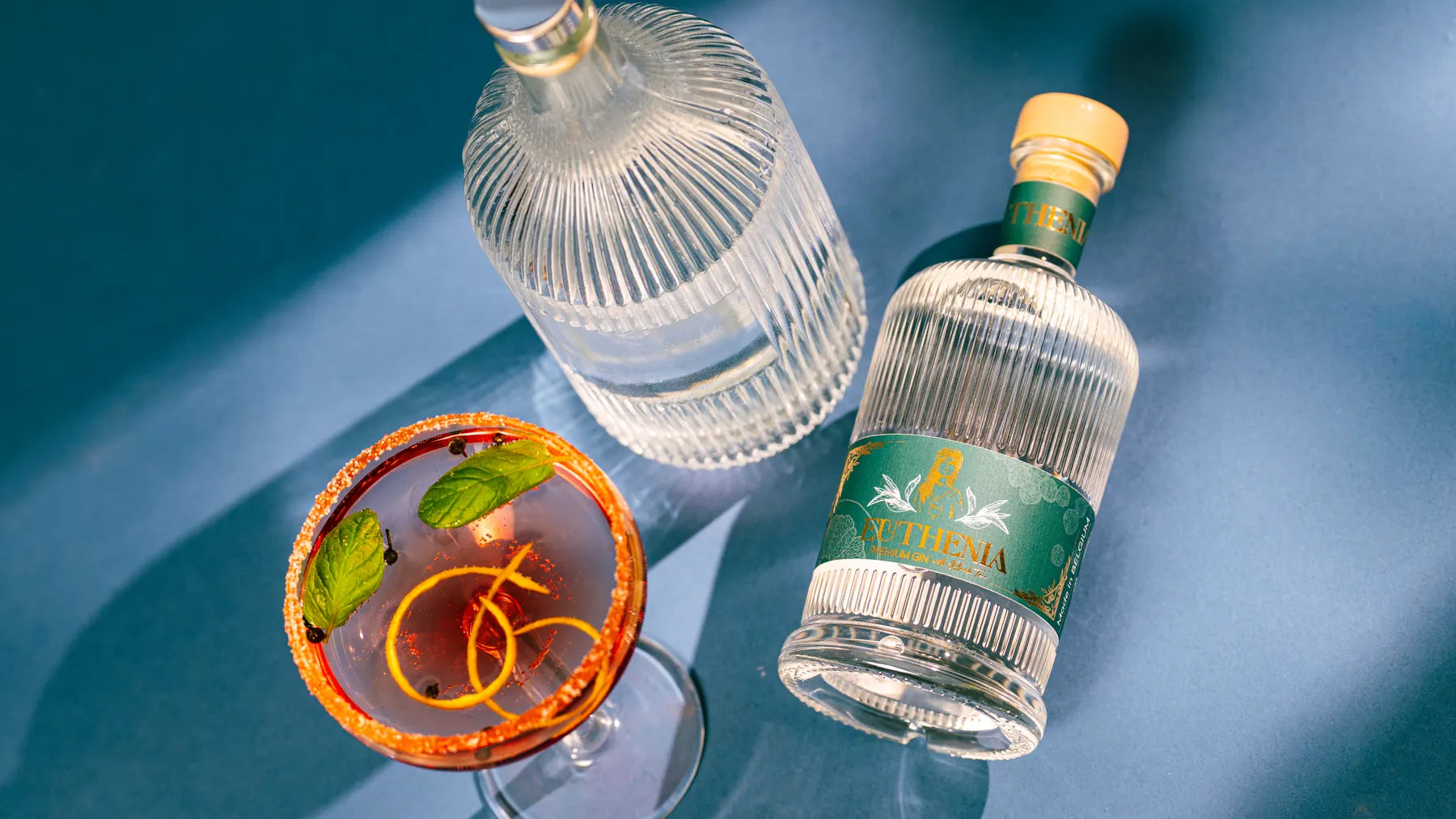 Bouteille de Gin Euthénia vue de haut, avec une coupe de cocktail orangée, menthe et zeste, sur fond bleu